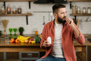 Young attractive man sitting at the kitchen table and taking essential Vitamins and Minerals for Adult, looking away. Healthy lifestyle concept.